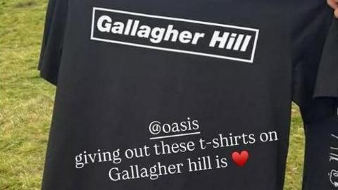 A picture of a black t-shirt with the words 'Gallagher Hill' in white lettering, with an Instagram caption reading '@Oasis giving out these t-shirts on Gallagher Hill is' followed by a heart emoji. 
