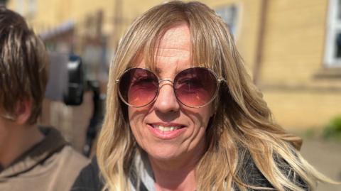 Janet is sitting down and is in the middle. She has long blonde hair and a fringe and is wearing sunglasses and a black coat.