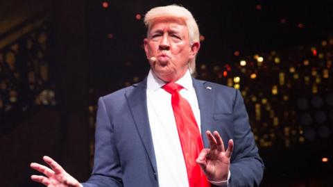 Mike Osman dressed as Donald Trump. He is wearing a blonde wig and pursing his lips together. He is wearing a blue suit and red tie.