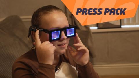 A girl wears a pair of glasses with thick black frames and lenses that are lit up with blue lights. She's pressing a button on the side of the glasses.