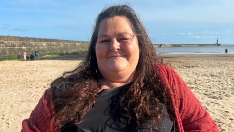 Katie Wotton smiling into the camera on a beach. She's got long, curly brown hair and is wearing a red cardigan with a black top.