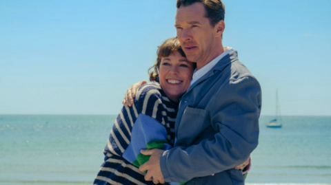 A man and a woman in an embrace in front of a beach. Calm waters lap onto the sandy shoreline behind them.