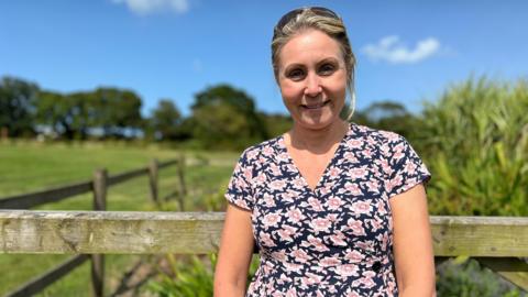 Denise smiles at the camera as she stands in front of a fence with a field in the background. She is wearing a dress with pink flowers on a black background. She has sunglasses on her head covering some of her blonde hair.