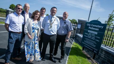 Six people, including the mayor Ruairí McHugh and a number of council officials, are standing beside the cemetery sign that reads: City Cemetery Mullenan. Black gates and a grassy embankment are visible in the background. 
