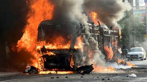 A bus is engulfed in flames, with thick black smoke billowing into the air as fire spreads around the vehicle.