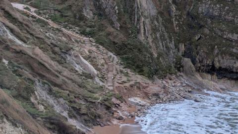 View from the top of the cliffs looking down at Durdle Door beach and the steep steps leading down to the sand. The bottom section of the steps appear to be missing along with a chunk of the cliff that has been washed away.