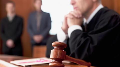 Stock photo of a judge and gavel in court. There's a shot of a slightly out of focus man dressed in black as a judge. There's two people standing in the background in suits.