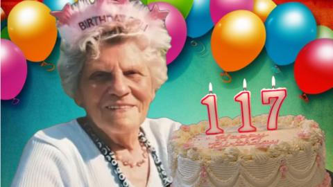An old woman in a cardigan and necklace sitting in front of a background with multi-coloured balloons wearing a hat that says Birthday Girl! on it. To the right is a cake with candles reading 117.