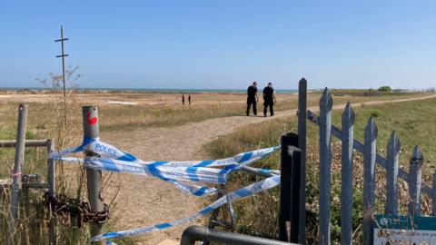 Police officers walk down a path towards the beach. The path is taped off with blue and white tape.