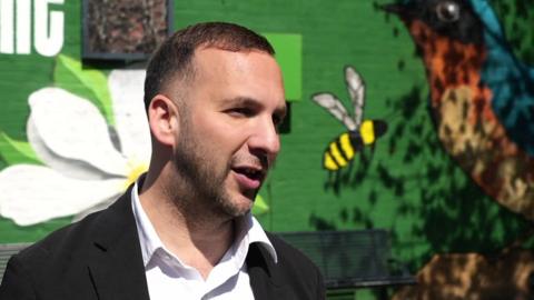 A man in a black blazer and white shirt looks towards a camera while standing in front of a dark green painted wall. 