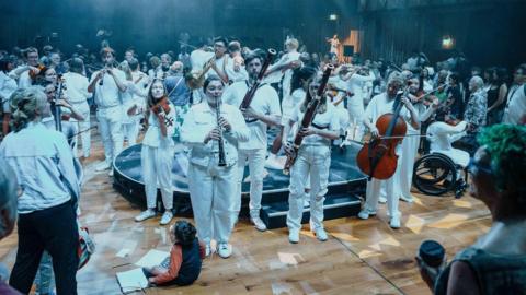 A goup of musicians in a circle, all wearing white, playing their instruments, surrounded by members of the audience