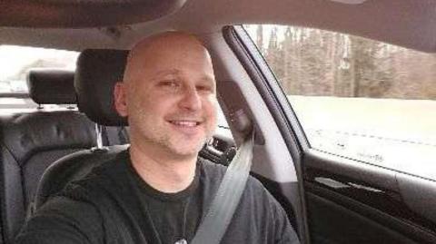 Man in a car. he is smiling and wears a seatbelt across his shoulder. He is bald and the weather looks snowy.