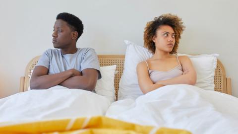 Couple lay in bed looking away from each other as if they're arguing.