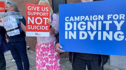 People holding up signs for and against the bill. The include a blue sign with the words Campaign for Dignity in Dying, and a red sign with Support Not Suicide written on them. There is also a waste paper bin with the words Bin the Bill on it.