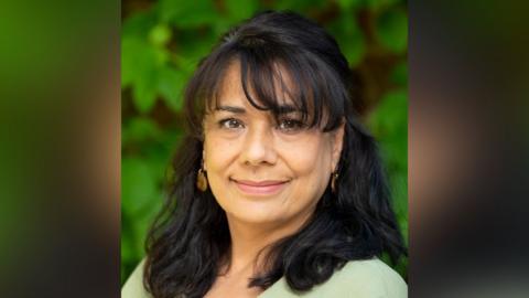 A woman with long black hair and earrings who is smiling facing the camera. She is wearing a light brown top.