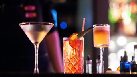 Three cocktails sit on a bar counter: a clear drink in a martini glass, a tall patterned glass with an orange‑garnished drink and a straw, and a pink‑orange cocktail with a foamy top, all set against a blurred backdrop of colourful bar lights.