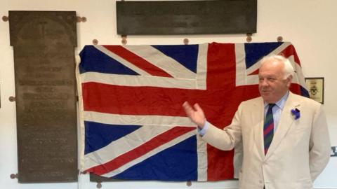 The Lord Lieutenant of Lincolnshire, Toby Dennis, wears a pale cream jacket and stands in front of a large Union flag at the unveiling of two bronze tablets containing the names of soldiers who died during the First World War.