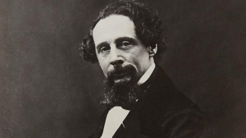 A black and white image of Charles Dickens he has curly balding hair and a goatee beared. He wears a black bow-tie and suit jacket.