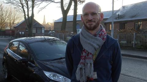 Gareth Cartwright is stood outside next to his black Ford Fiesta car. He has a bald head and is wearing a grey scarf and blue zipped jumper.