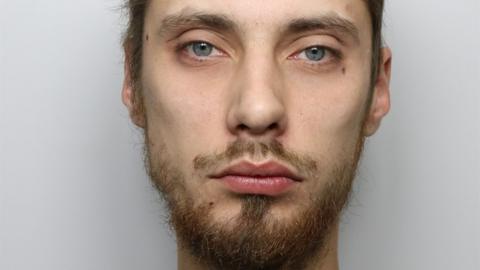 A man with short brown hair and brown or ginger facial hair and blue eyes. It is a custody photo and the background is grey.