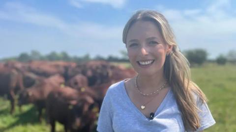 A smiling Martha Hayes standing in front of a herd of Lincoln Reds wearing a grey top and necklace.