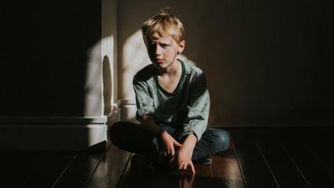 A young boy sat alone on a wooden floor, staring sadly at the camera