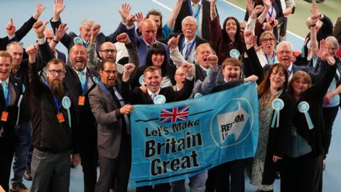 A group of Reform UK members at the election count in Sunderland. They are cheering and holding a blue banner that reads, Let's Make Britain Great.