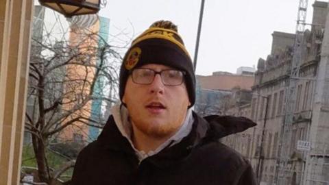 A man in his mid-20s with red hair and a short beard, wearing glasses and a dark hat