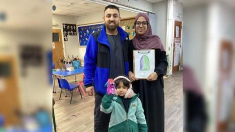 Akeel, Hannaa and Sara in Al Ashraf Primary School. Sara is wearing a warm coat, pink earmuffs and has a hand puppet. Hannaa is holding a drawing done by Sara.