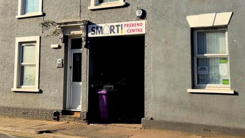 The outside of a day centre, showing a grey building with white windows, a purple rubbish bin, a sign above a gap and a path in front. An alarm is on the wall and a doorway is to the left, with a tree growing out of the top of a lintel.