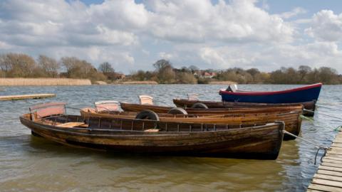 Boats moored on the water at Hornsea Mere. There are trees in the background and the sky is blue with fluffy cloud.