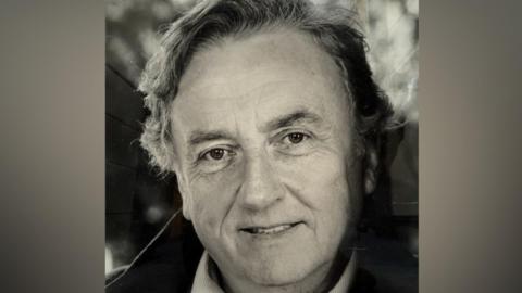 A black and white head and shoulders image of a man smiling at the camera. He has grey hair and is wearing a light-coloured shirt.