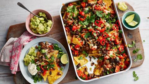 A baking dish filled with tortilla chips, mince, sliced chillis and sour cream. A scoop has been taken out and put on a blue plate beside it. Side bowls of guacamole and lime wedges also sit on a chopping board.