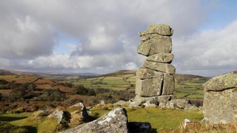 Nine giant boulders are piled on top of each other on a grassy outcrop. There are singular boulders lying on the ground nearby. All of them are covered in lichen. Dartmoor stretches out behind them, covered with fields, hedges and bracken. It is sunny but there are also clouds. 