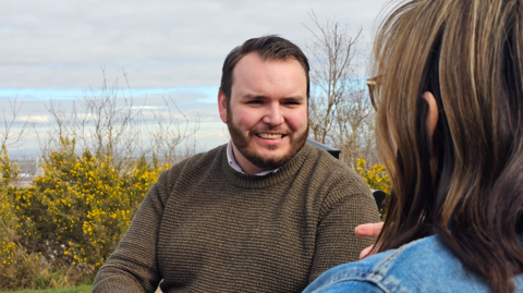 Sean Houlston smiling in a dark green jumper with an unknown woman in the foreground