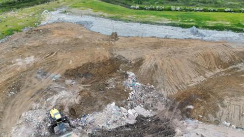 Drone image of the landfill site taken from above showing a vast area of brown mud, surrounded by green fields. There is a yellow tractor in the middle of the mud with debris and piles of rubbish.