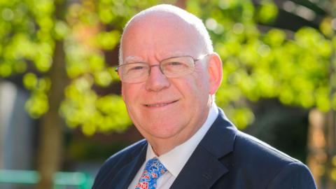 A head-and-shoulders shot of a smiling bald man wearing glasses and a suit with a flowery tie, with a tree in soft focus behind him.