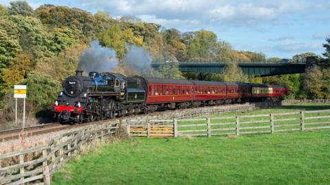 The Pocket Rocket is a black locomotive. Grey smoke puffs out of its funnel and trails behind it over claret coloured carriages. The locomotive is passing through an area of countryside with trees on one side of the track and fields on the other. It is a sunny day.