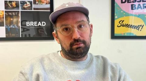 A man wearing a lilac cap, round, clear glasses, and a grey jumper. He has a short beard, and is looking into the camera. Behind him there are framed signs.
