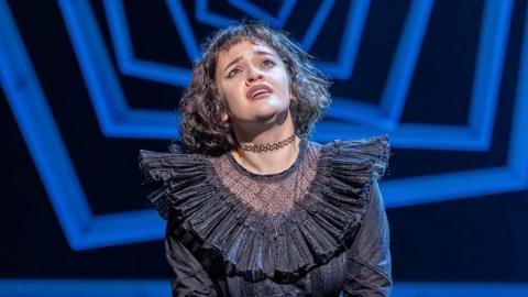 Rachel is wearing a frilly black costume, and a choker around her neck. She has shoulder length black hair and is looking up into the sky.