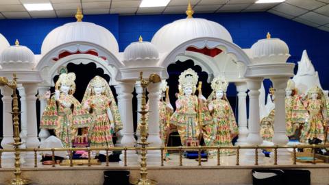 Seven Hindu marble statues wearing gold, green and pink robes under a white dome. 