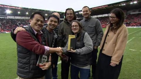 A group of people are stood on a football pitch, with a woman in the middle accepting a black ands gold coloured plaque and shaking hands with a man on the left hand side of the photo.