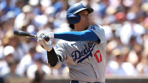 Shohei Ohtani in a Dodgers jersey on the baseball field, swinging his bat with both arms. His eyes are slightly open and he is wearing a blue helmet. 