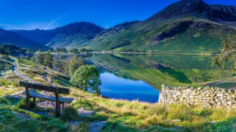A wooden bench in the foreground overlooks Buttermere. Hills in the distance are reflected in the lake on a sunny day.