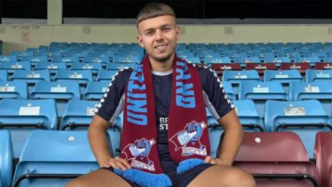 Jonathan Gjoshe sitting in the stands at Glanford park wearing blue shorts and shirt and a Scunthorpe United scarf