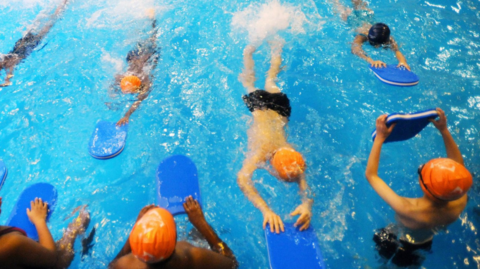Children swimming in a leisure centre pool carrying floats wearing goggles and swim caps