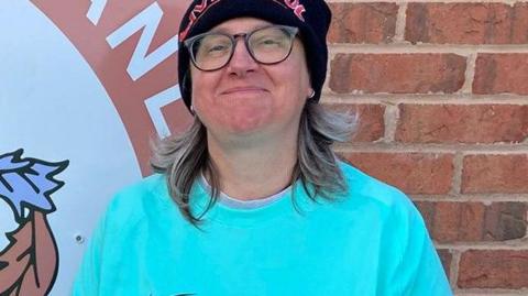 Eileen Fox, who has shoulder-length dark blonde hair, is wearing glasses, a turquoise t-shirt and a blue woollen hat with 'Liverpool' stitched across the hem in red letters. The 56-year-old is smiling for the camera while standing in front of a brick wall.