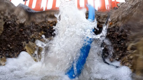 Water is gushing into a hole in a road. The image is close up from inside the hole and looking upward. There is a blue pipe half in and half out of the hole. Above on the road, you can see orange fencing around the hole.