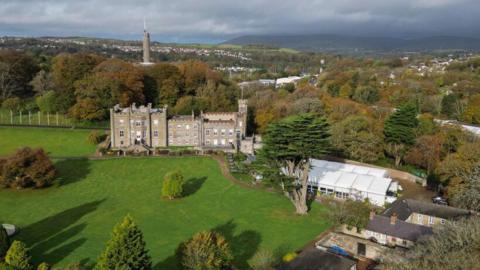 A large historic stone mansion sits in the middle of sweeping green lawns and trees. The estate is a small castle set within a private park. Beyond the grounds are autumn-coloured woodlands and a small town 