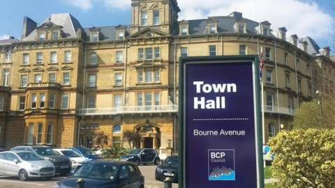 Bournemouth Town Hall is a historical building in light brown stone, at least five storeys high. In front is a car park and purple sign carrying the BCP Council logo.